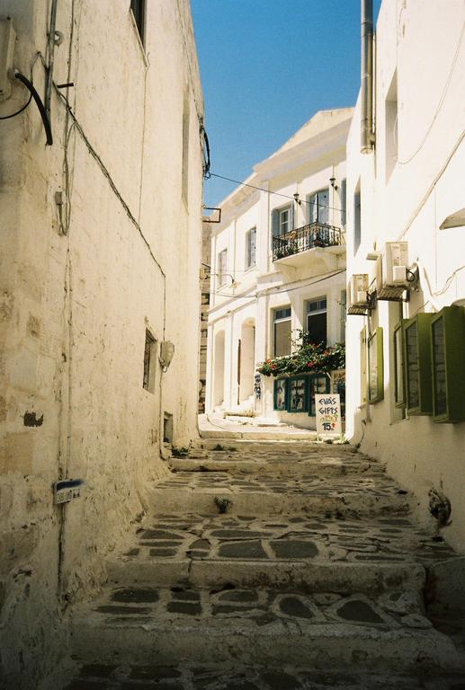 A narrow, stepped alleyway with whitewashed buildings on either side leads up to a distant building with a balcony and green shutters.