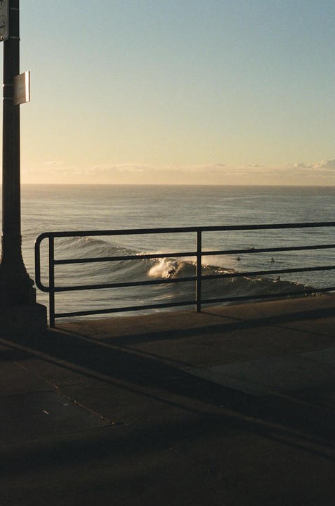 A surfer rides a wave in the ocean, viewed from behind a metal railing on a pier.