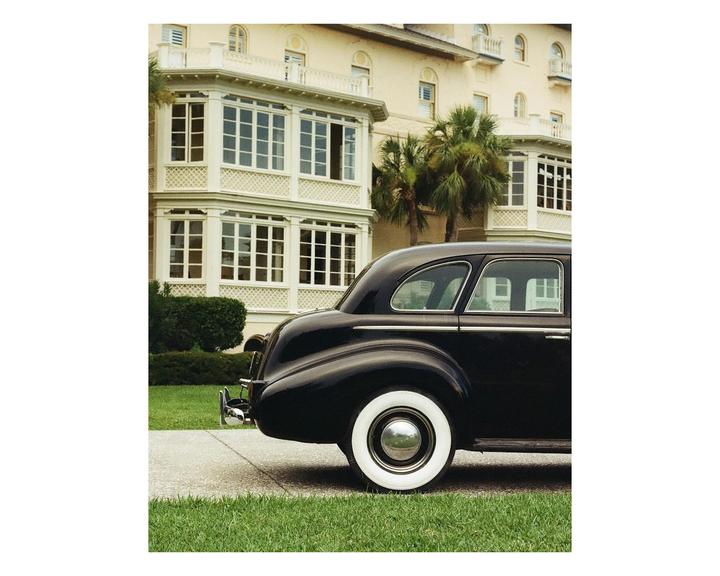 A black vintage car with whitewall tires is parked on a driveway in front of a large building with palm trees.