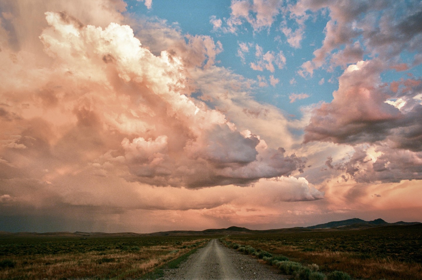 A dirt road leads through a vast, flat landscape under a dramatic sky filled with pink and orange clouds at sunset.