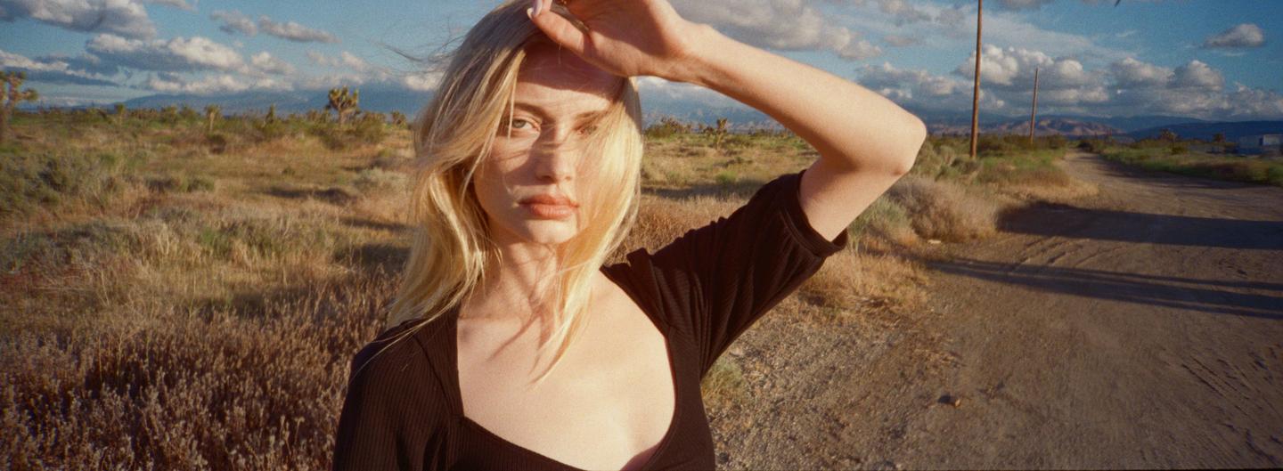 A woman shields her eyes from the sun in a desert landscape with a dirt road and mountains in the distance.