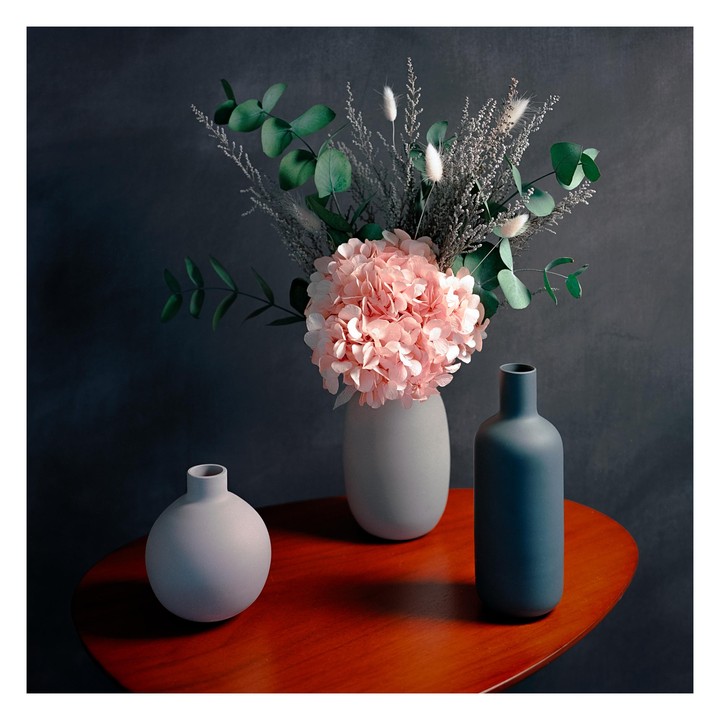 A still life composition features a vase of pink flowers and greenery, flanked by two smaller vases, all resting on a polished wooden table.