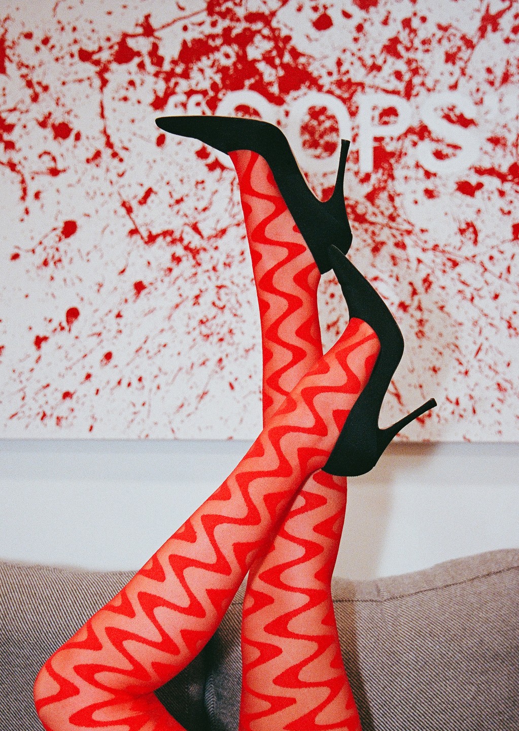 A person's legs in red patterned tights and black high heels are propped up against a couch with a red and white abstract painting in the background.