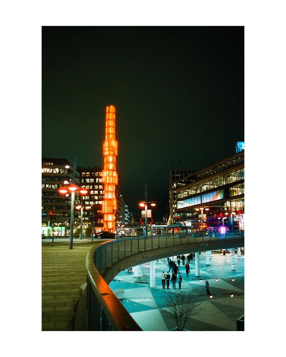 A tall, illuminated orange tower stands among city buildings at night, with people walking on a plaza below a curved pedestrian bridge.