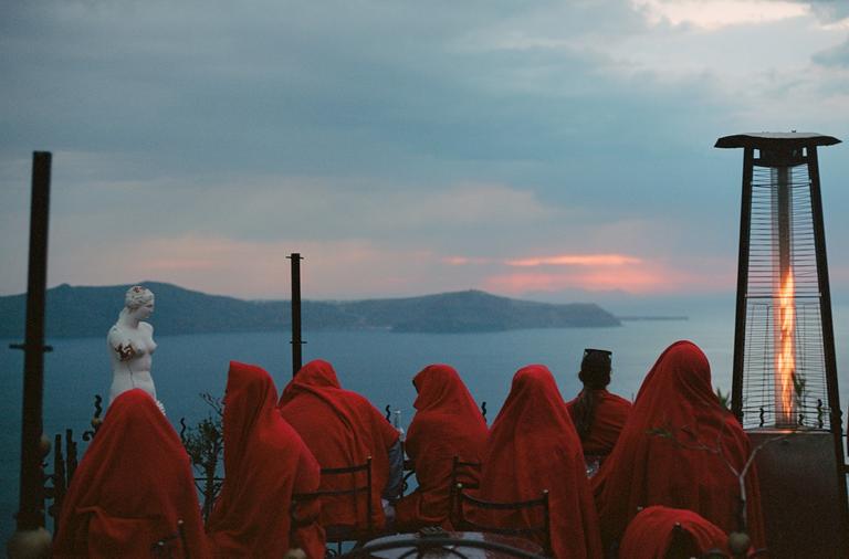 People wrapped in red blankets sit at tables overlooking a body of water with islands and a sunset.