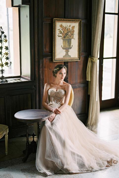 A woman in a long, flowing gown sits at a small round table next to a window.