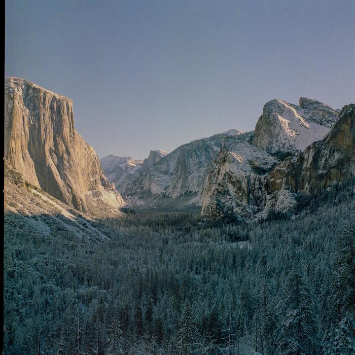A valley filled with snow-covered trees is surrounded by large, rocky mountains, with one mountain face illuminated by sunlight.
