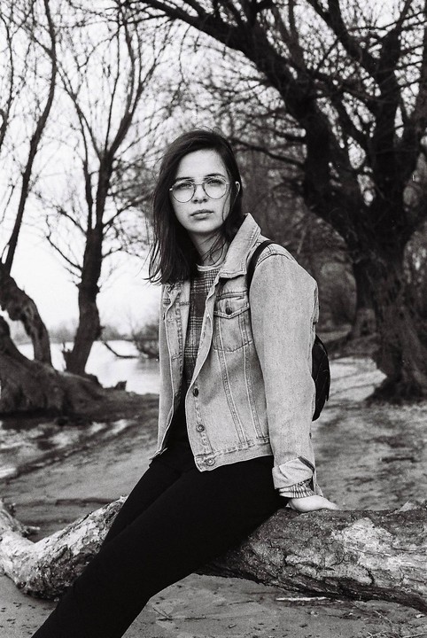 A woman wearing glasses and a denim jacket sits on a fallen tree trunk near a body of water with bare trees in the background.