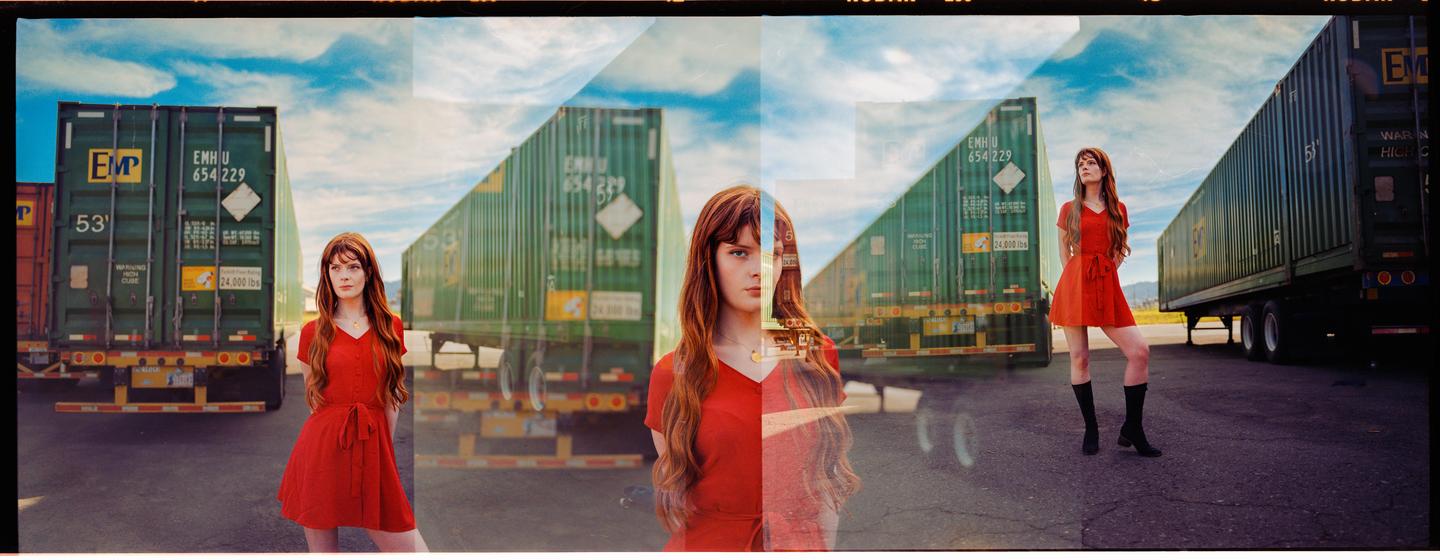 A woman in a red dress is triple exposed in front of green shipping containers under a blue sky.