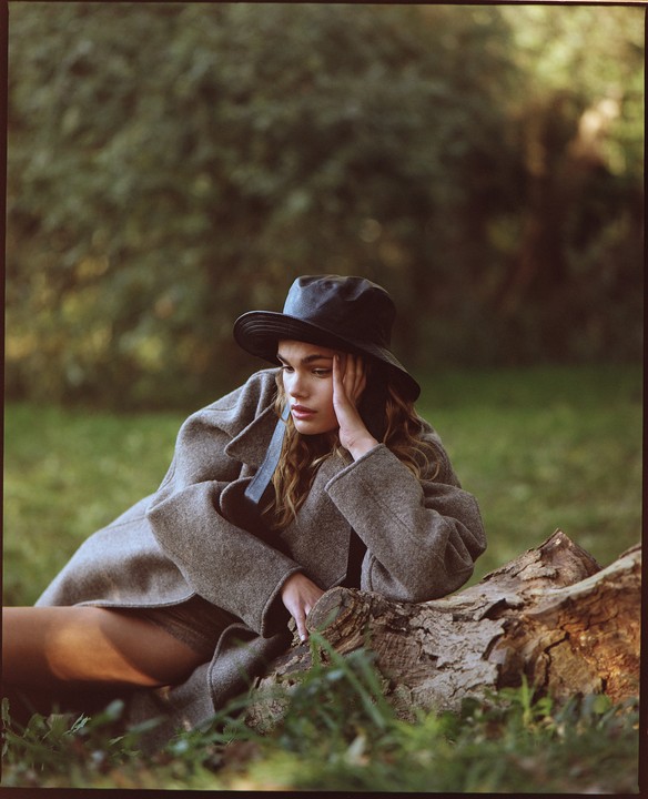 A woman in a hat and coat rests her arm on a log in a grassy area.