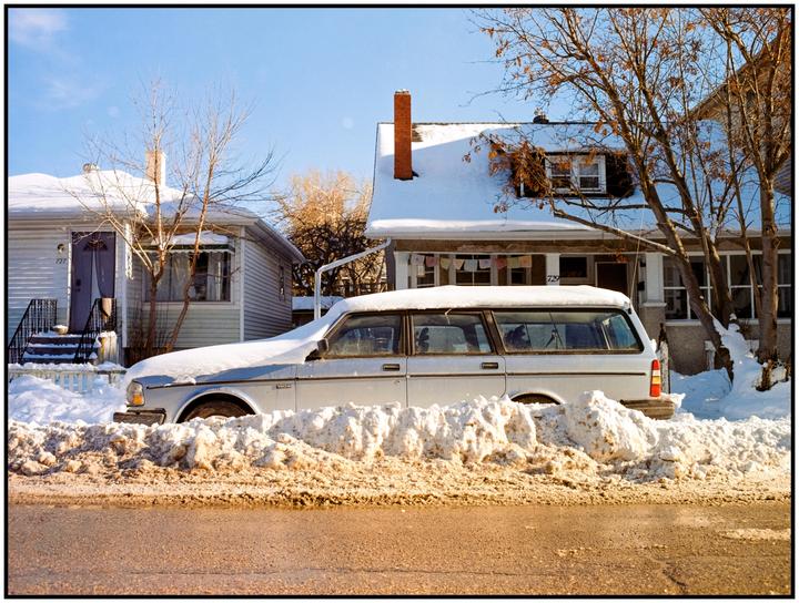 A snow-covered station wagon is parked on a street in front of two houses with snow on their roofs.