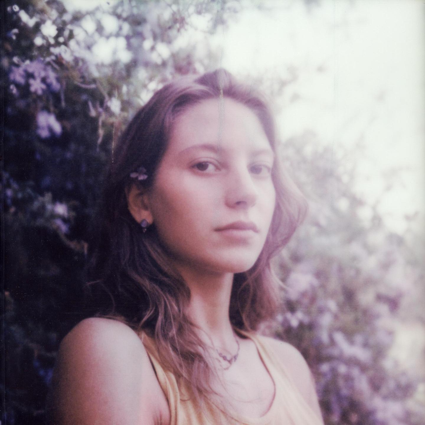 A woman with long hair and a flower behind her ear looks directly at the viewer.