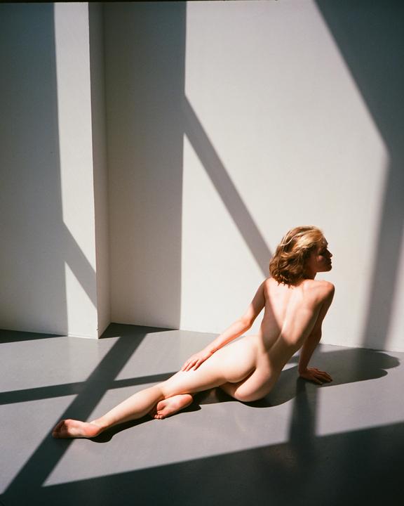A nude woman reclines on a floor with strong shadows cast across the wall and floor.