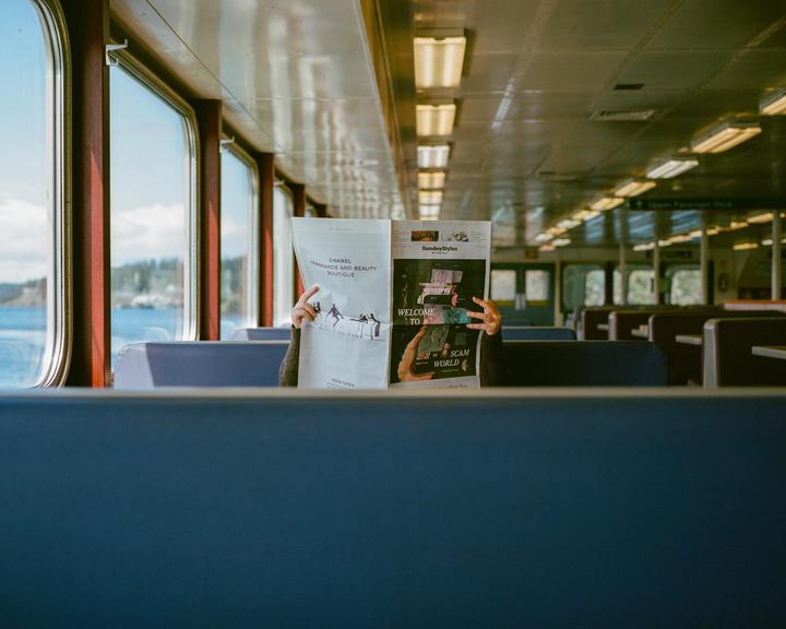 A person reads a newspaper on a ferry with water and land visible through the windows.