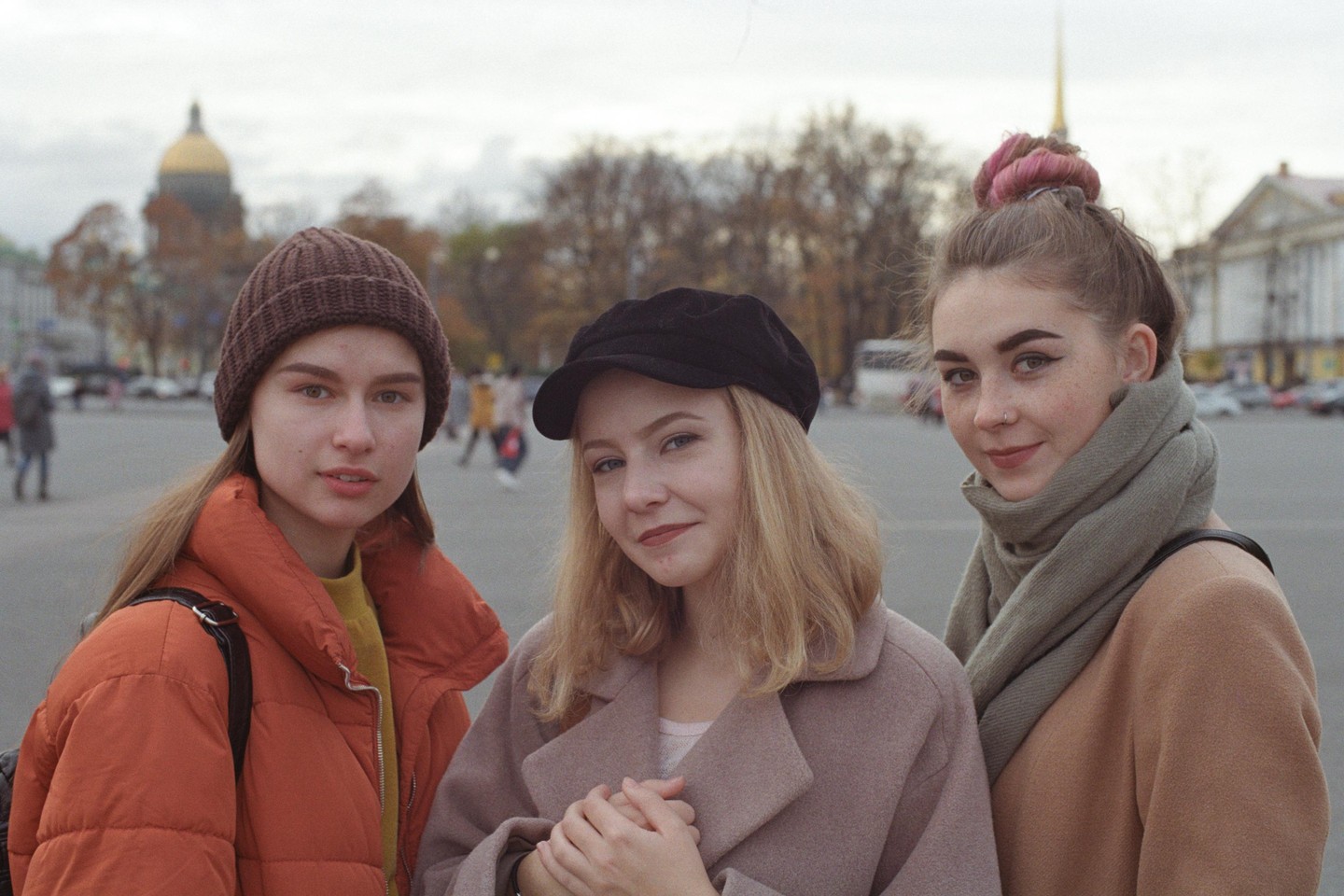 Three young women pose for a portrait on a city street with a golden domed building in the background.