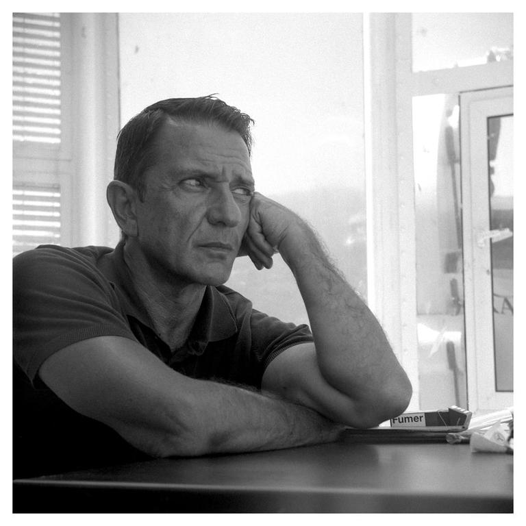 A man with his hand on his face looks away from the camera while sitting at a table.