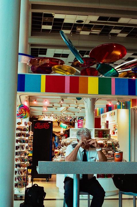 A man in a pilot uniform sits at a counter in a colorful shop with his head in his hands.