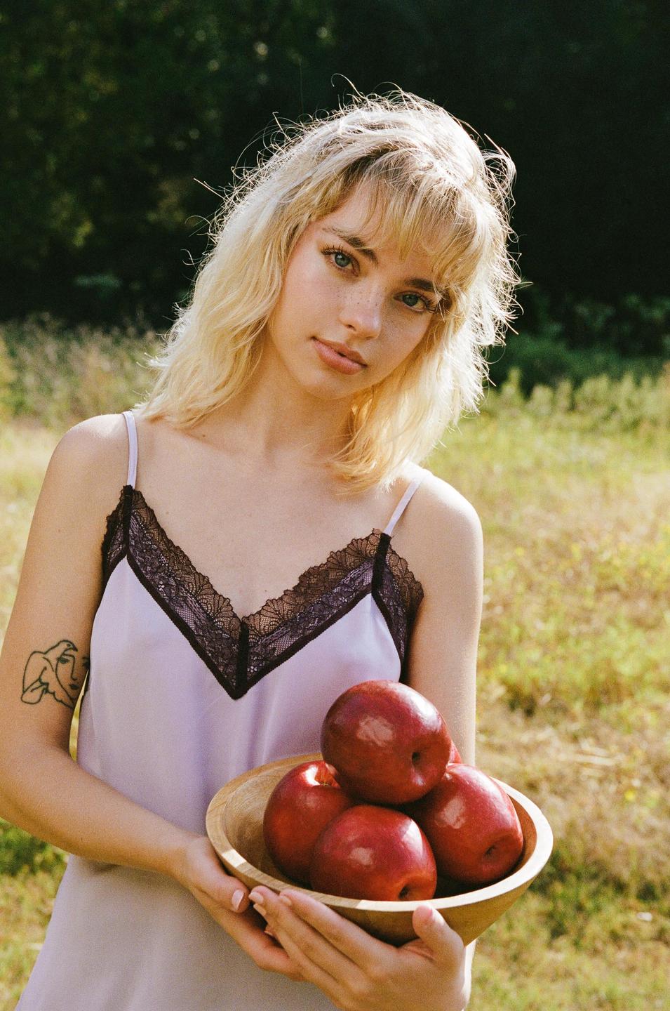 A woman with blonde hair holds a wooden bowl of red apples in a grassy field.