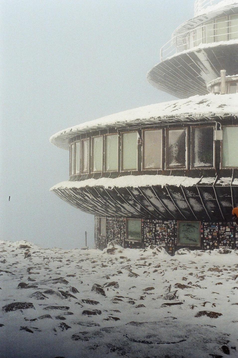 A snow-covered building with many windows stands on a rocky, snowy landscape under a hazy sky.