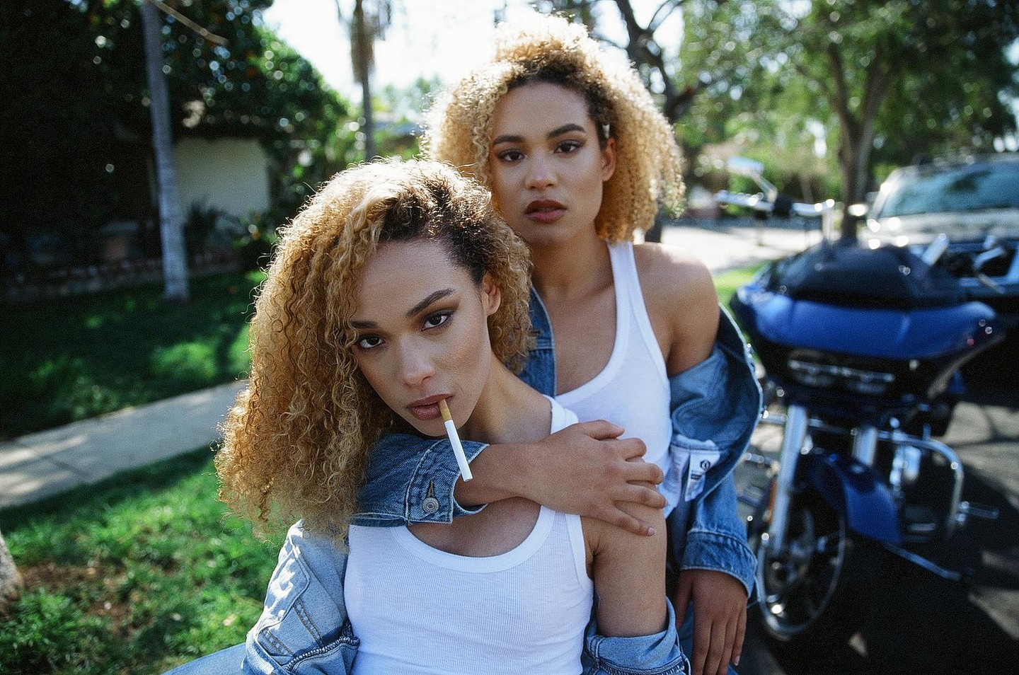 Two women with curly hair pose for a portrait, one with a cigarette in her mouth and the other with her arm around her.