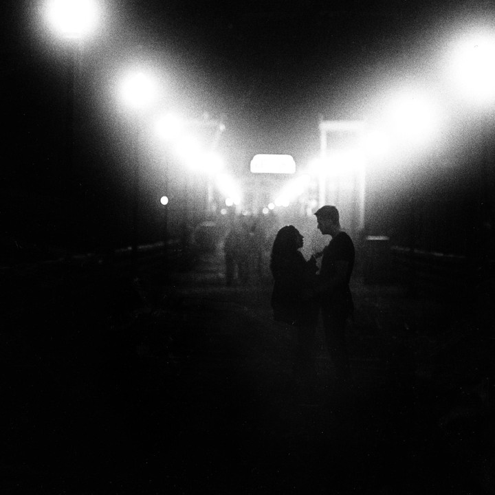 A couple stands silhouetted against bright lights at night.