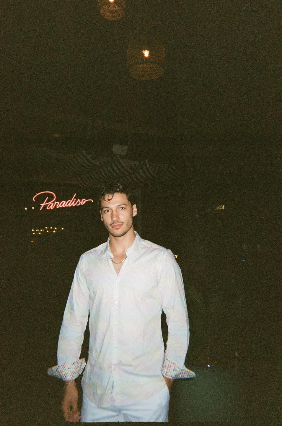 A man in a white shirt stands in front of a neon sign that reads "Paradiso" at night.