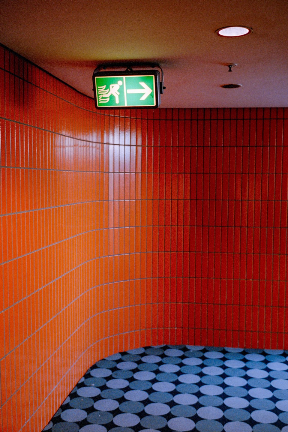 A green exit sign hangs from a ceiling above a curved wall of orange and red tiles and a patterned floor.