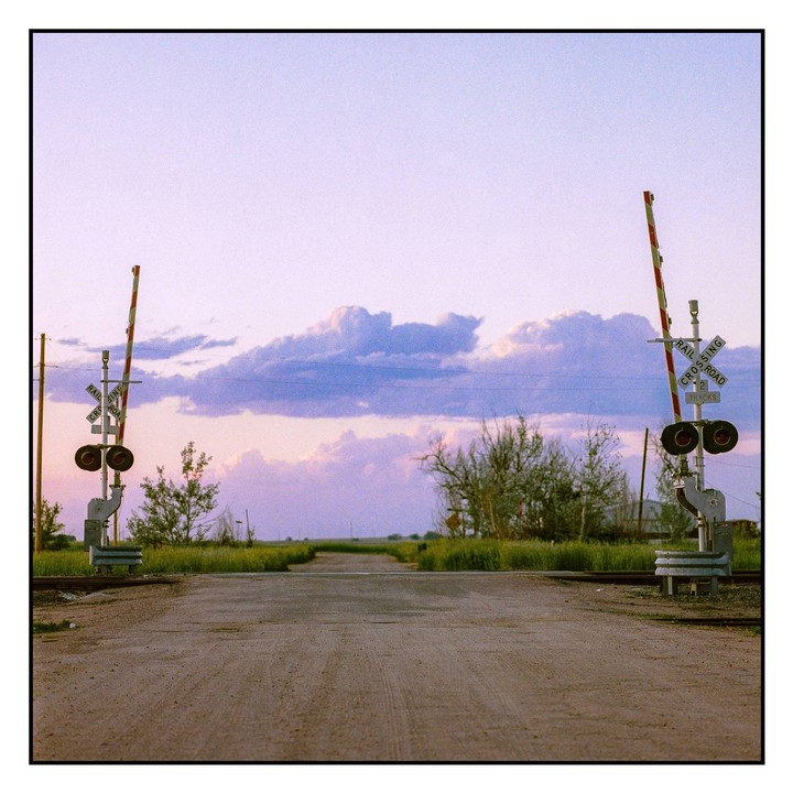 A dirt road leads to a railroad crossing with a vast sky above.