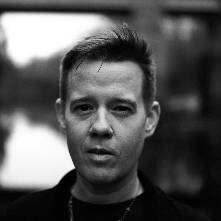 A man with short hair and a necklace looks directly at the viewer.