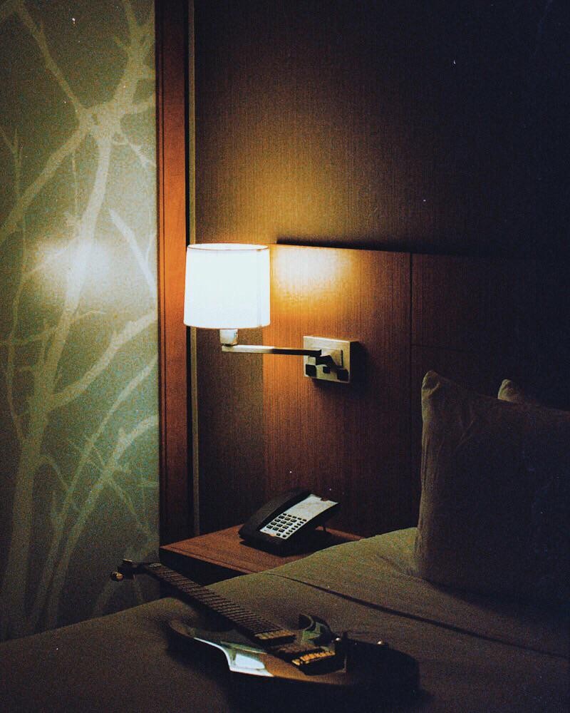 An electric guitar rests on a bed next to a nightstand with a telephone and a lit lamp in a hotel room.