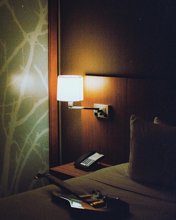 An electric guitar rests on a bed next to a nightstand with a telephone and a lit lamp in a hotel room.