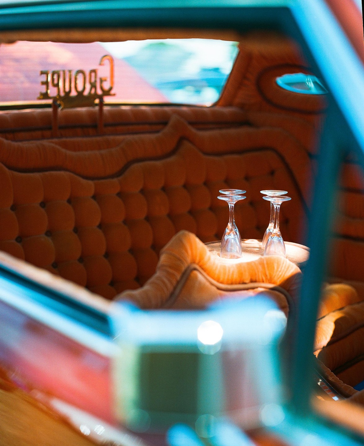 Two champagne flutes sit on a small table in the back of a car with plush orange upholstery.