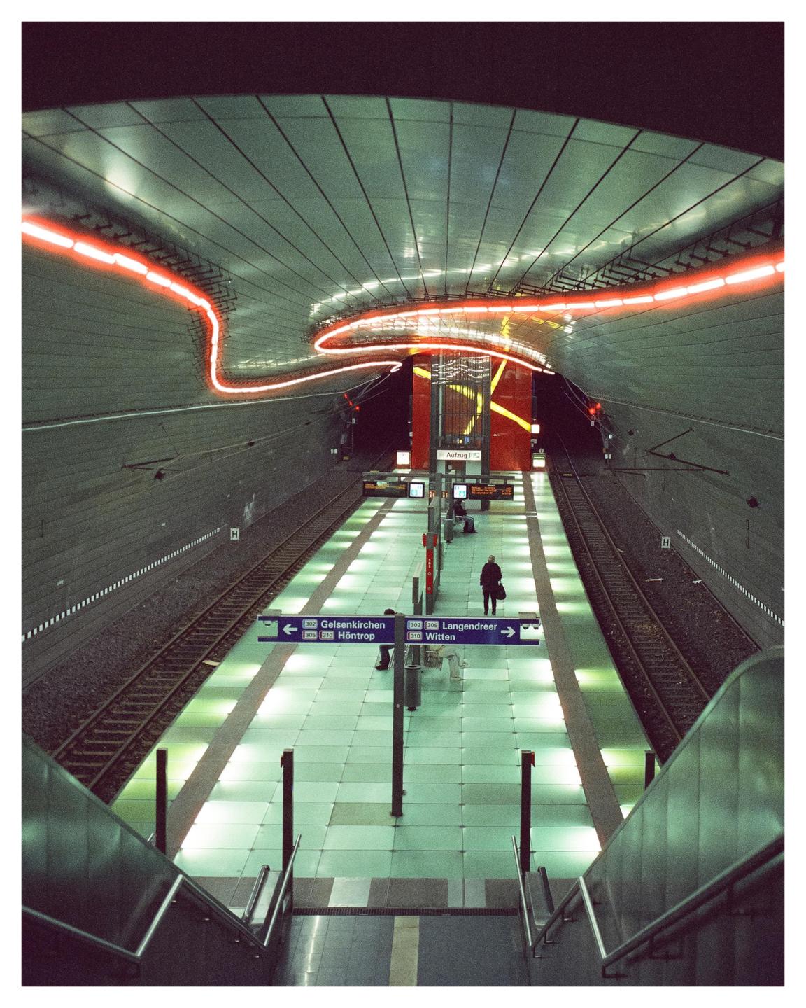 A person stands on a subway platform with light trails from a passing train overhead.