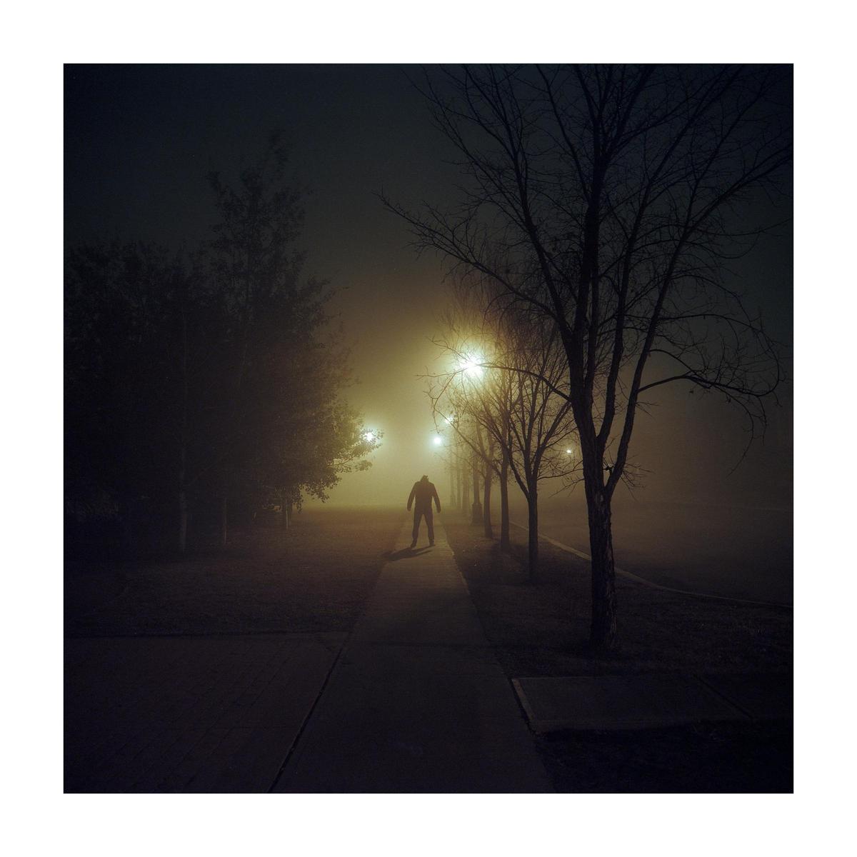 A lone figure stands on a sidewalk at night, illuminated by streetlights in a thick fog.