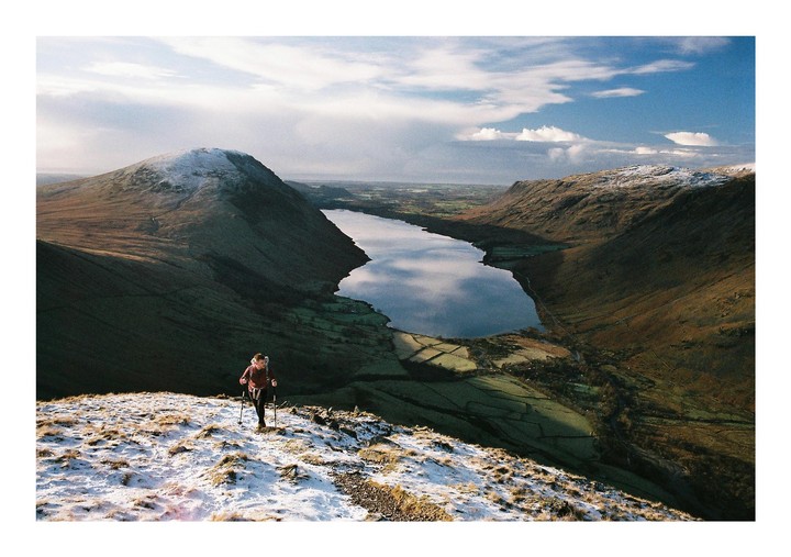 A person hikes up a snowy mountain path overlooking a lake surrounded by fells.