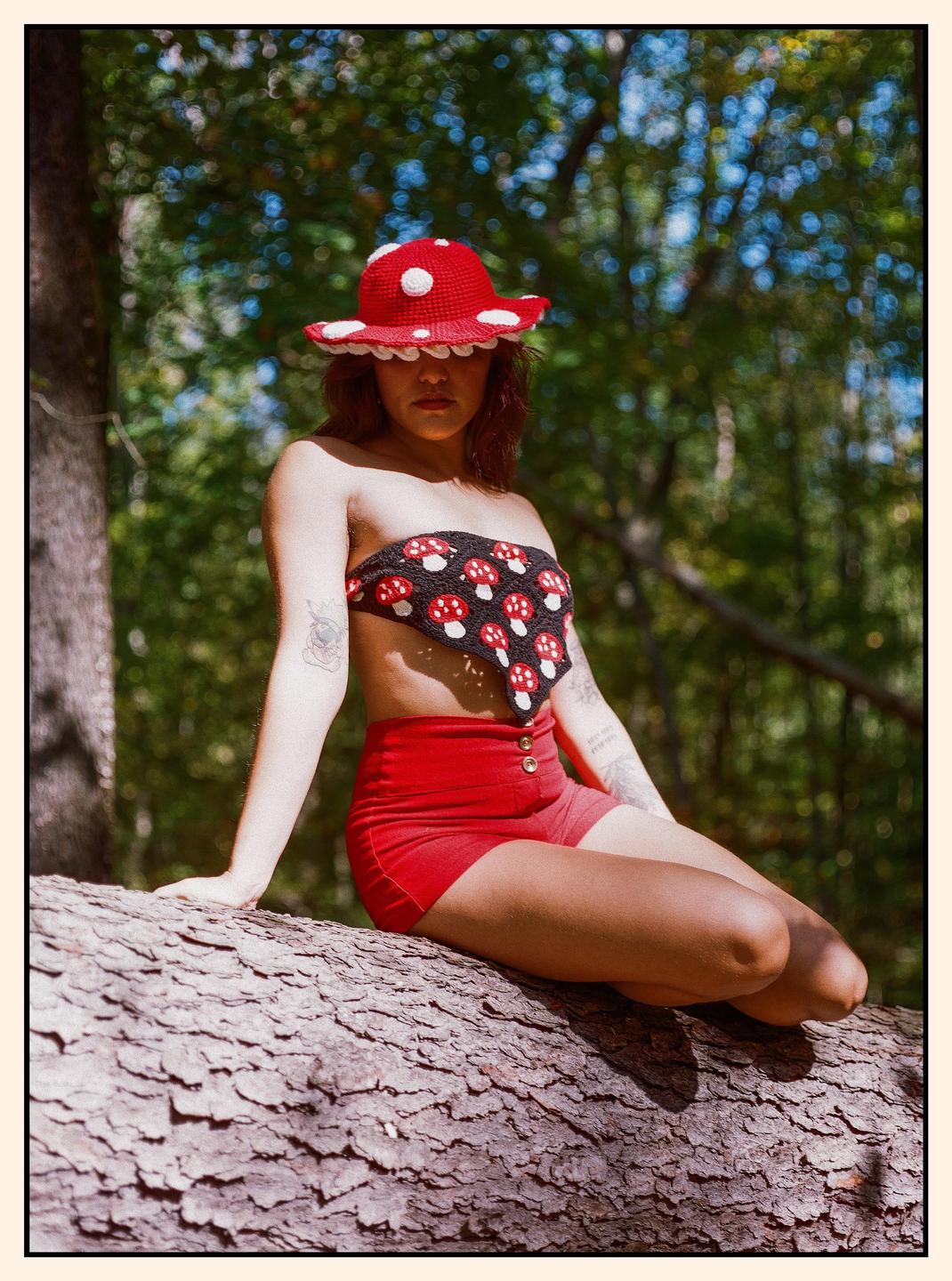 A woman wearing a mushroom hat and top sits on a log in a forest.