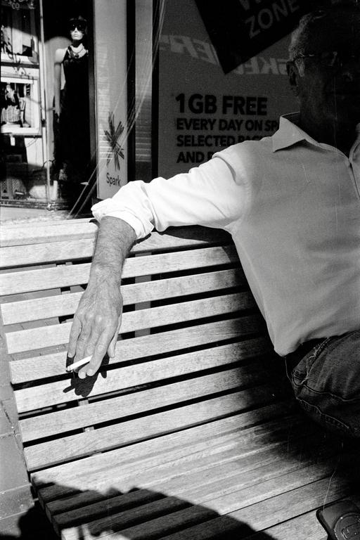 A man sits on a wooden bench with a cigarette in his hand, with a store window and mannequin visible in the background.