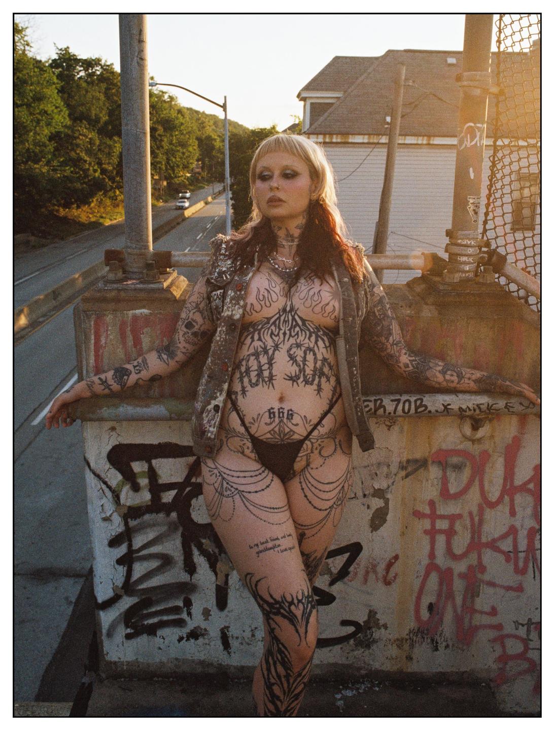 A woman with extensive tattoos poses on a graffiti-covered ledge overlooking a road at golden hour.