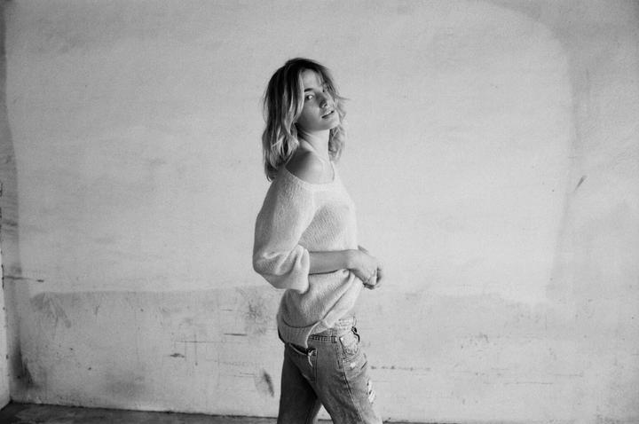 A woman wearing a sweater and jeans stands against a textured wall, looking over her shoulder.