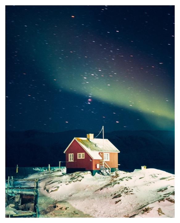 A red house sits on a snowy hill under a starry night sky with the aurora borealis.