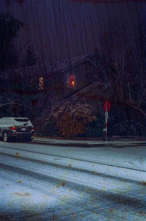 A car is parked on a snowy street at night, with a house and trees in the background.
