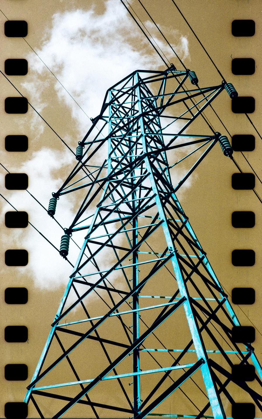 A large metal electricity pylon stands against a sky with clouds, with film sprockets visible on the sides.