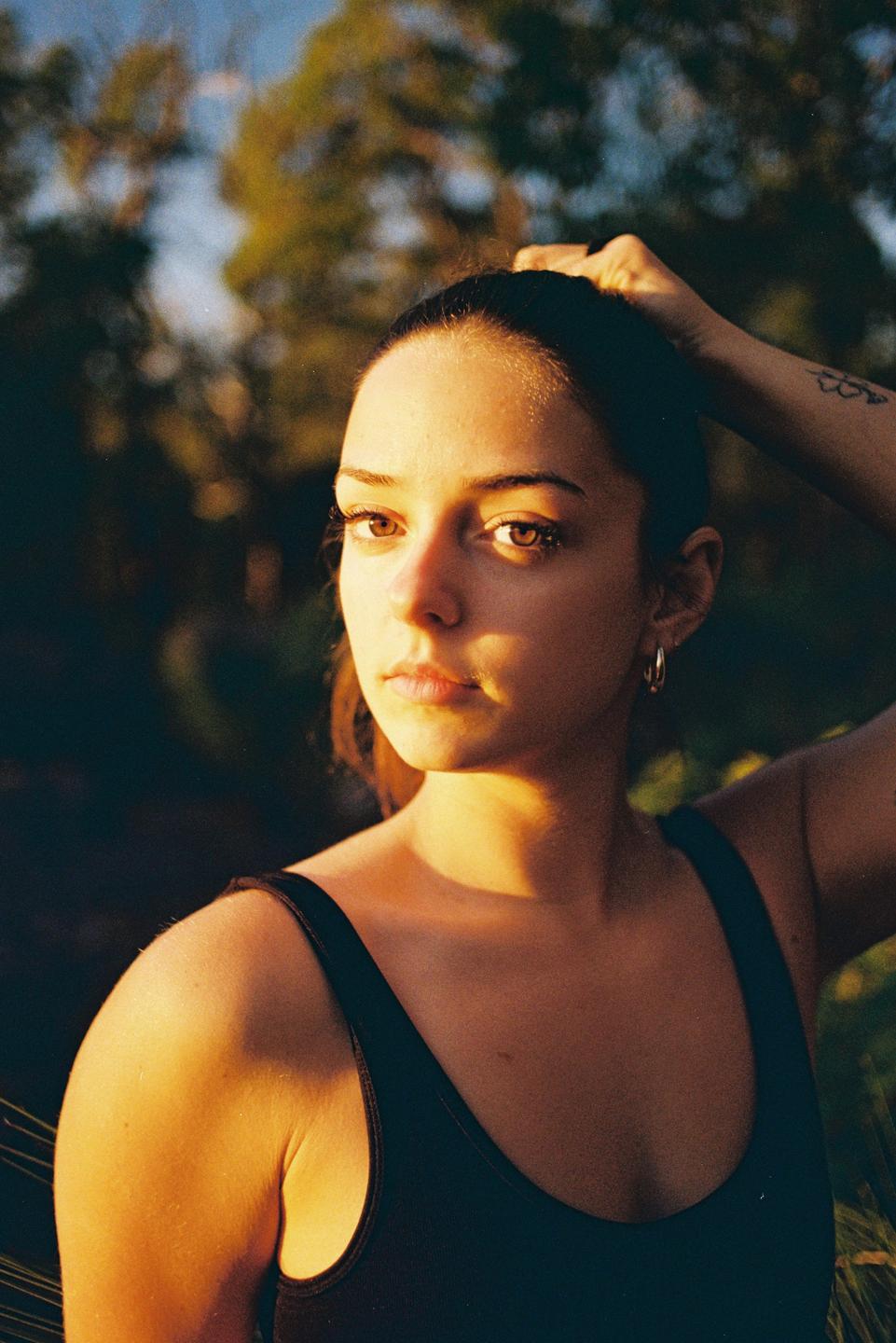 A woman with dark hair pulled back looks directly at the viewer, with golden light illuminating her face and shoulder.