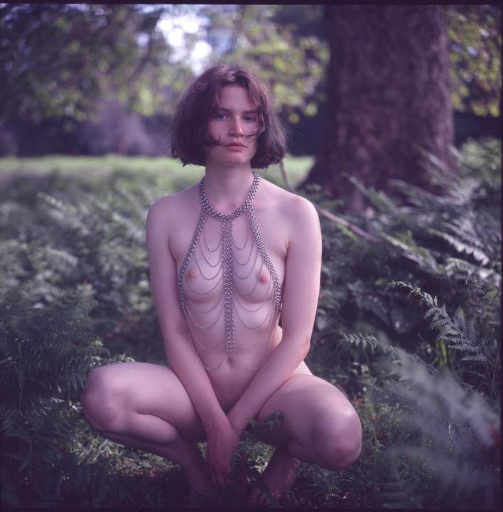 A woman wearing a chain top crouches in a forest.