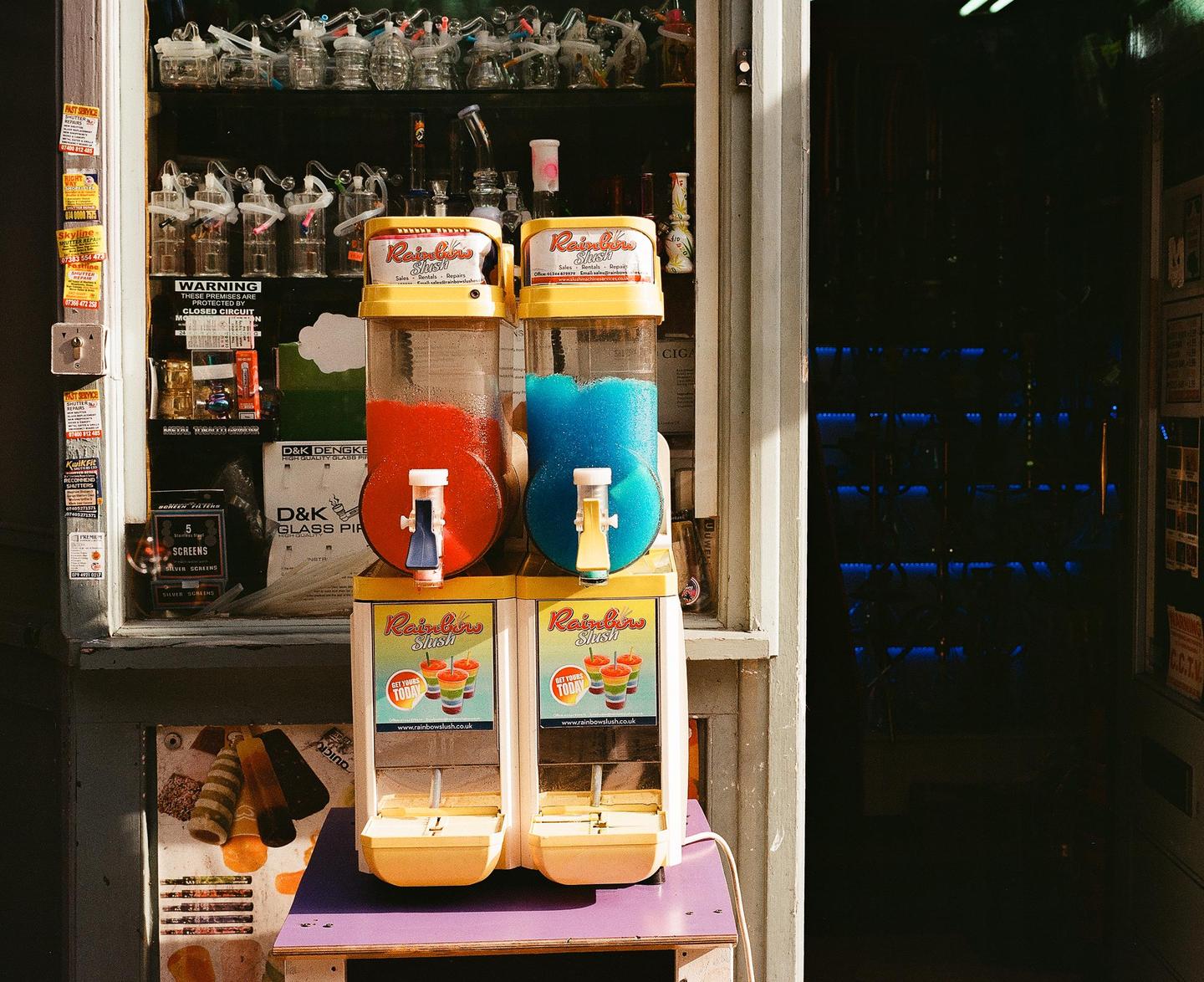 Two slushie machines with red and blue drinks sit on a table outside a shop.
