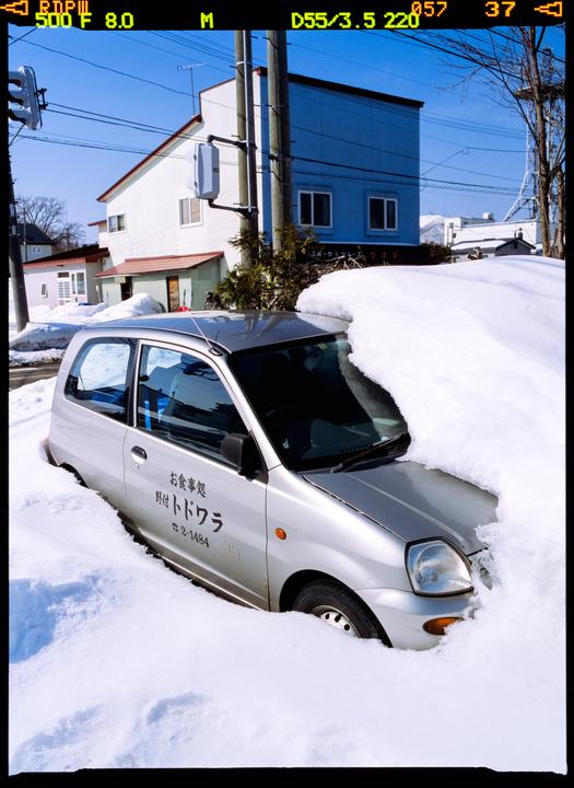 A silver car is partially buried in deep snow on a street with buildings and utility poles in the background.