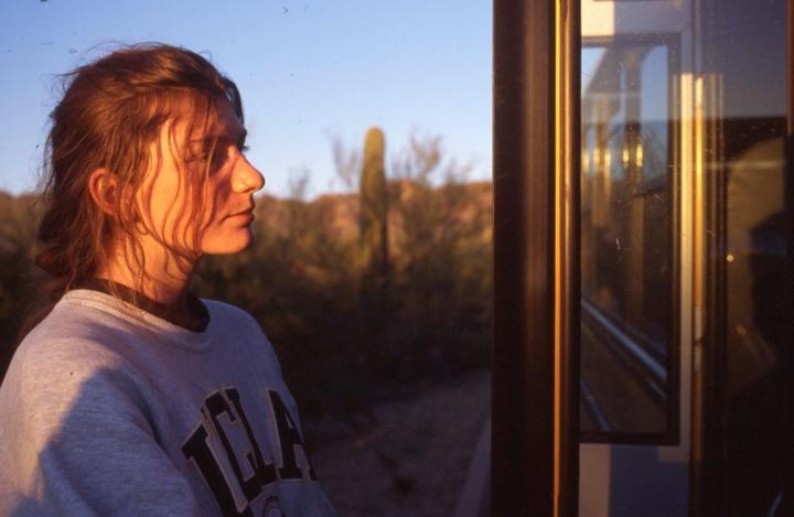 A woman with long hair looks to the right, with a desert landscape and a window visible in the background.