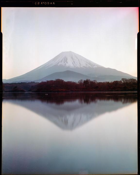 A snow-capped mountain is reflected in a still body of water, with a line of trees visible between the mountain and the water.