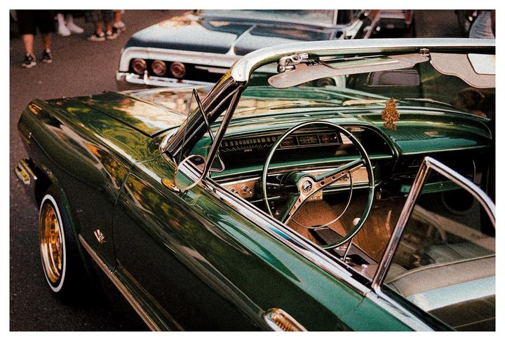 A green convertible lowrider is parked on a street with another car and people in the background.