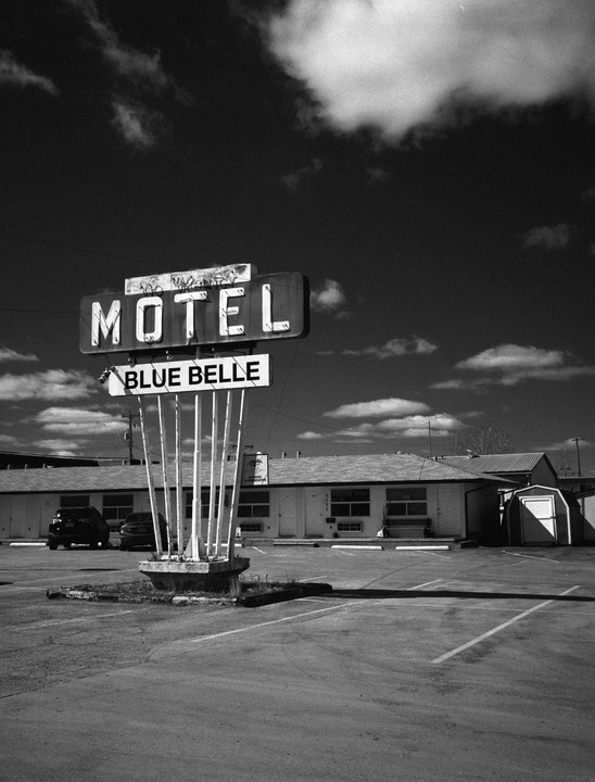 A black and white photograph shows the "Motel Blue Belle" sign in front of a motel building and parking lot under a cloudy sky.